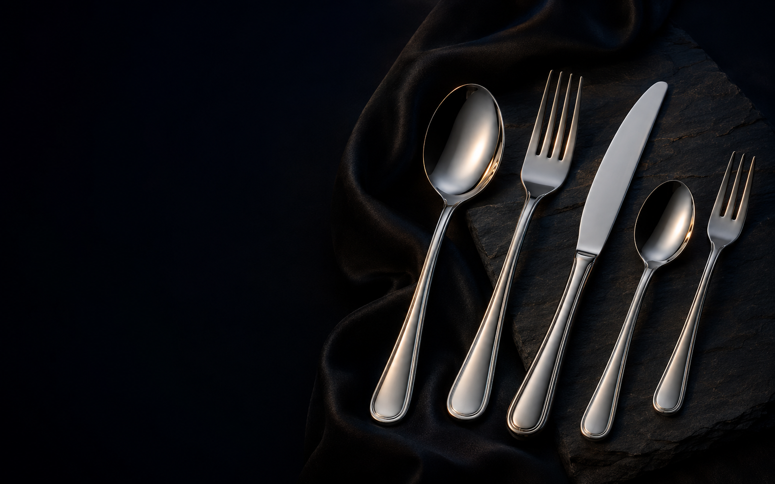 Polished flatware arranged on charcoal stone and black silk